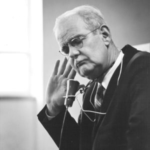 White man wearing glasses holding his hand to his ear with listening device around his neck