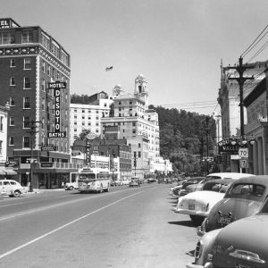 City street lined with multistory buildings, including "DeSoto hotel," and cars with wooded hillside in background