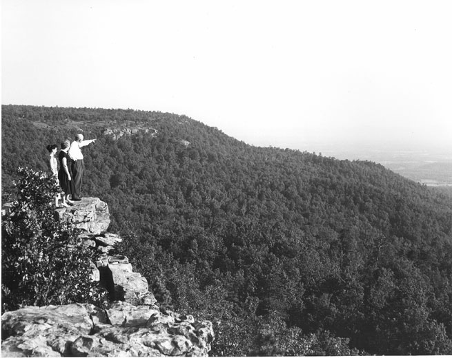 White man and two women standing on a cliff with mountain in the distance