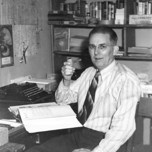 White man with glasses holding a pipe posing with an open book on his desk and bookshelves behind him