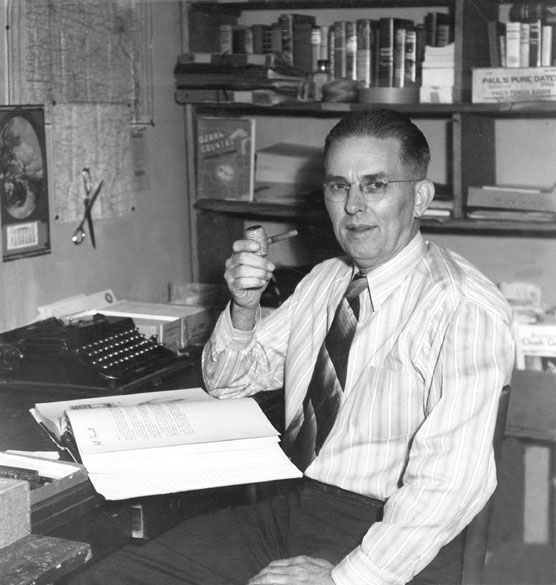 White man with glasses holding a pipe posing with an open book on his desk and bookshelves behind him