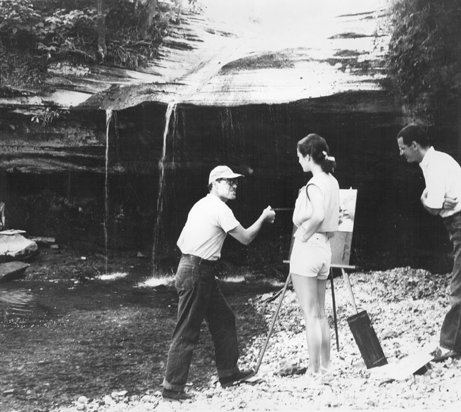 White man in cap and jeans talking to white man and woman while painting near a natural waterfall