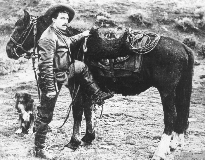 White man with mustache and cowboy hat mounting his horse while his dog stands nearby