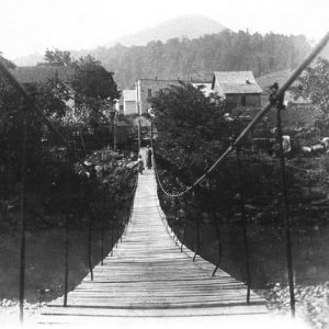 Man and woman on wooden suspension bridge over waterway with trees on either side and buildings in the distance