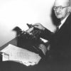 Old white man wearing glasses and a suit using a typewriter with group of papers next to him on the table