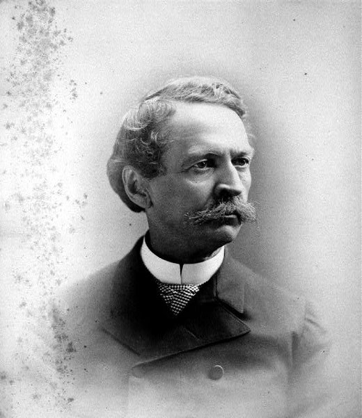 White man with curly hair and mustache in suit and white collar
