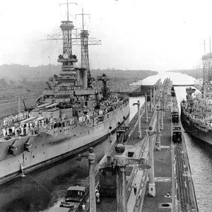 Two battleships covered in sailors in canal harbor with long dock including railroad tracks