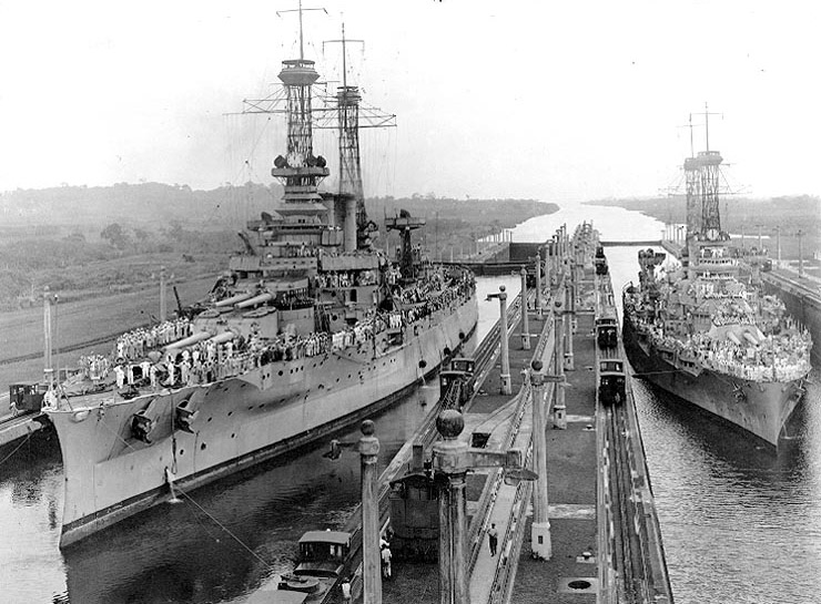 Two battleships covered in sailors in canal harbor with long dock including railroad tracks