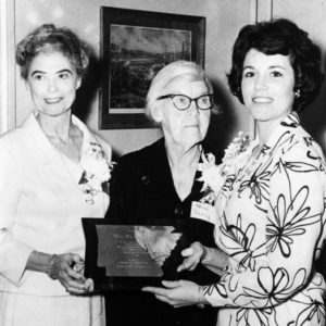 Three women formally dressed wearing corsages smile pose together holding Arkansas plaque
