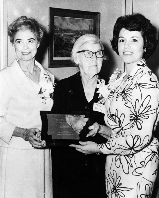 Three women formally dressed wearing corsages smile pose together holding Arkansas plaque