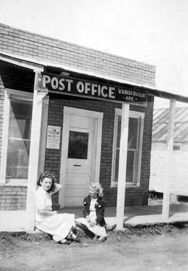 young white woman and blond girl sitting on the platform in front of a small brick building with a white door