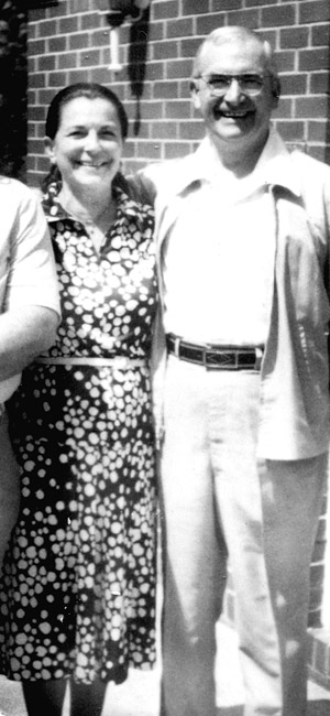 Smiling white woman in polka dot dress posing with white man in casual suit and glasses