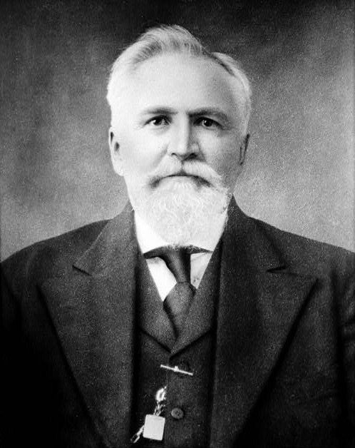 Portrait of an older white man with a white beard and receding hairline wearing a suit and tie
