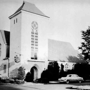 Multistory church with windowed tower arched entrance and hanging metal sign at street corner