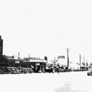Two-story lumber supply store with Chevrolet dealership and other stores on street with cars and power poles