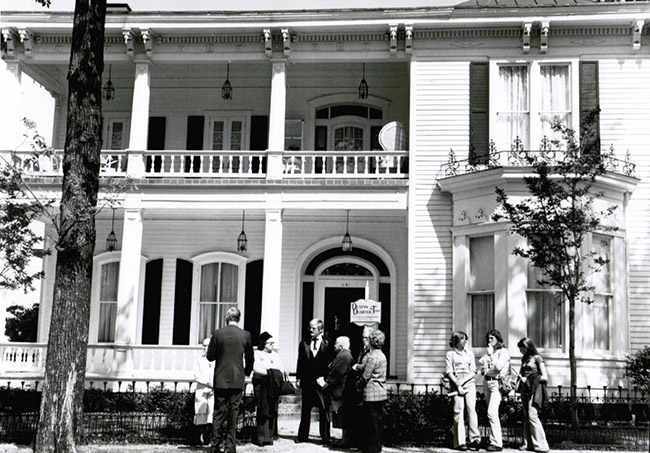 Group of white men and women standing outside multistory house with covered porch and balcony