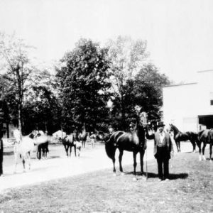 man in suit and top hat with horses and other men and stable building and trees