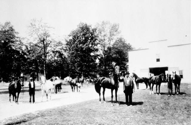 man in suit and top hat with horses and other men and stable building and trees