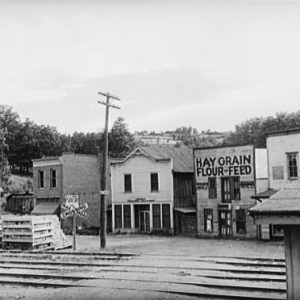 City street with wood frame buildings "Hay Grain Flour Feed" sign and railroad crossing
