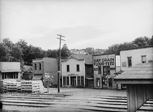 City street with wood frame buildings "Hay Grain Flour Feed" sign and railroad crossing