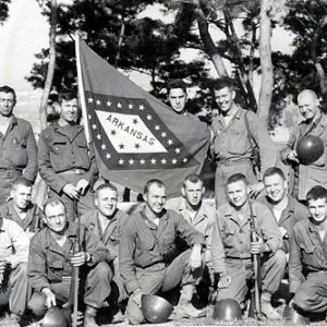 Group of men in military uniforms posing outdoors with Arkansas flag