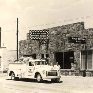 White man and truck in front of stone single-story building with sign saying "Alma Farm Supply Purina Chows"