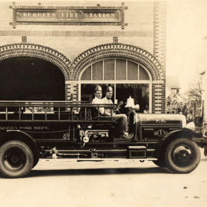 Two men sitting in fire truck in front of fire station