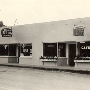 single story white brick building with "Corner Drug" and "Cafe" signs