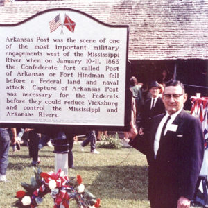 White man in suit and glasses standing next to historical marker and wreath