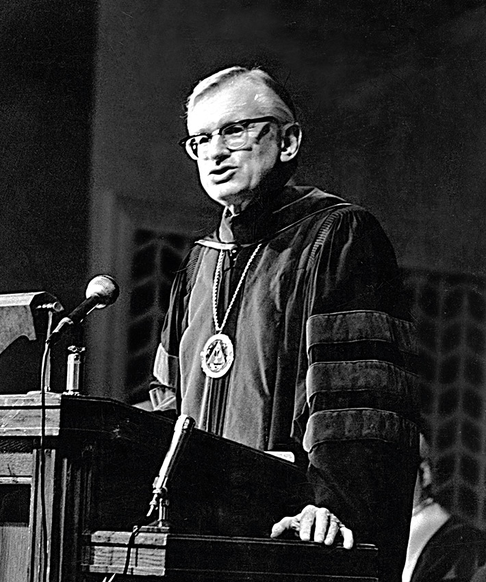 Man in graduation gown speaking at ;lectern