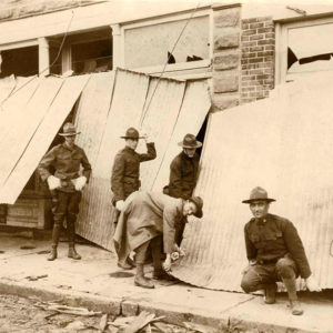 White men in military uniforms in front of damaged building removing sheet metal