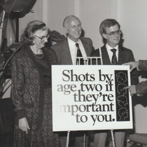 White woman and three white men behind sign promoting immunization.