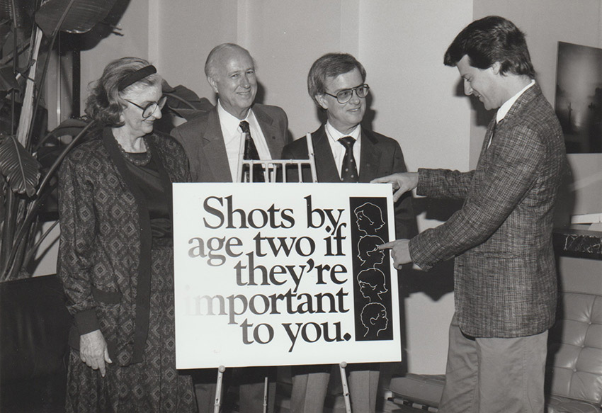 White woman and three white men behind sign promoting immunization.