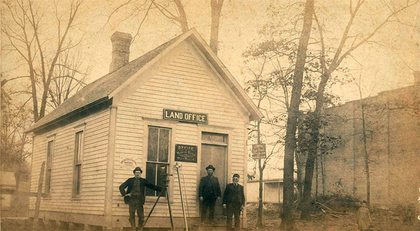 Three white men standing in front of white wooden building with sign saying "Land Office"