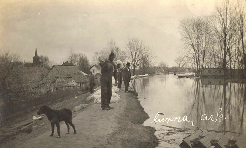 Men and dog standing on levee placing sandbags with houses and other buildings in the background