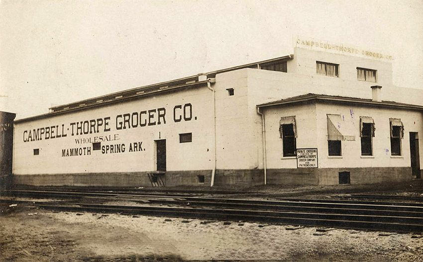 One story white building along railroad tracks with "Campbell-Thorpe Grocer Co." painted on the side
