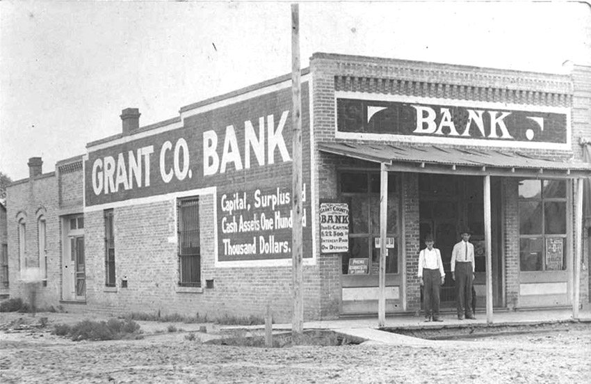 Two white men standing on porch of brick building with sign saying "Grant County Bank"