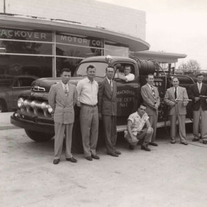 Group of white men standing around and sitting in fire truck parked before glass-fronted building