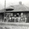 Group of people standing on porch of single story building next to railroad tracks