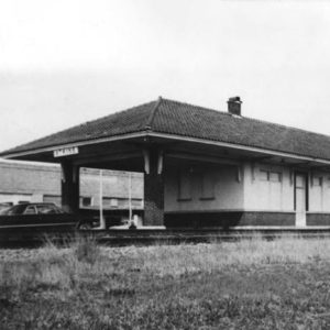 Single story building with brick columns and car in front beside railroad tracks