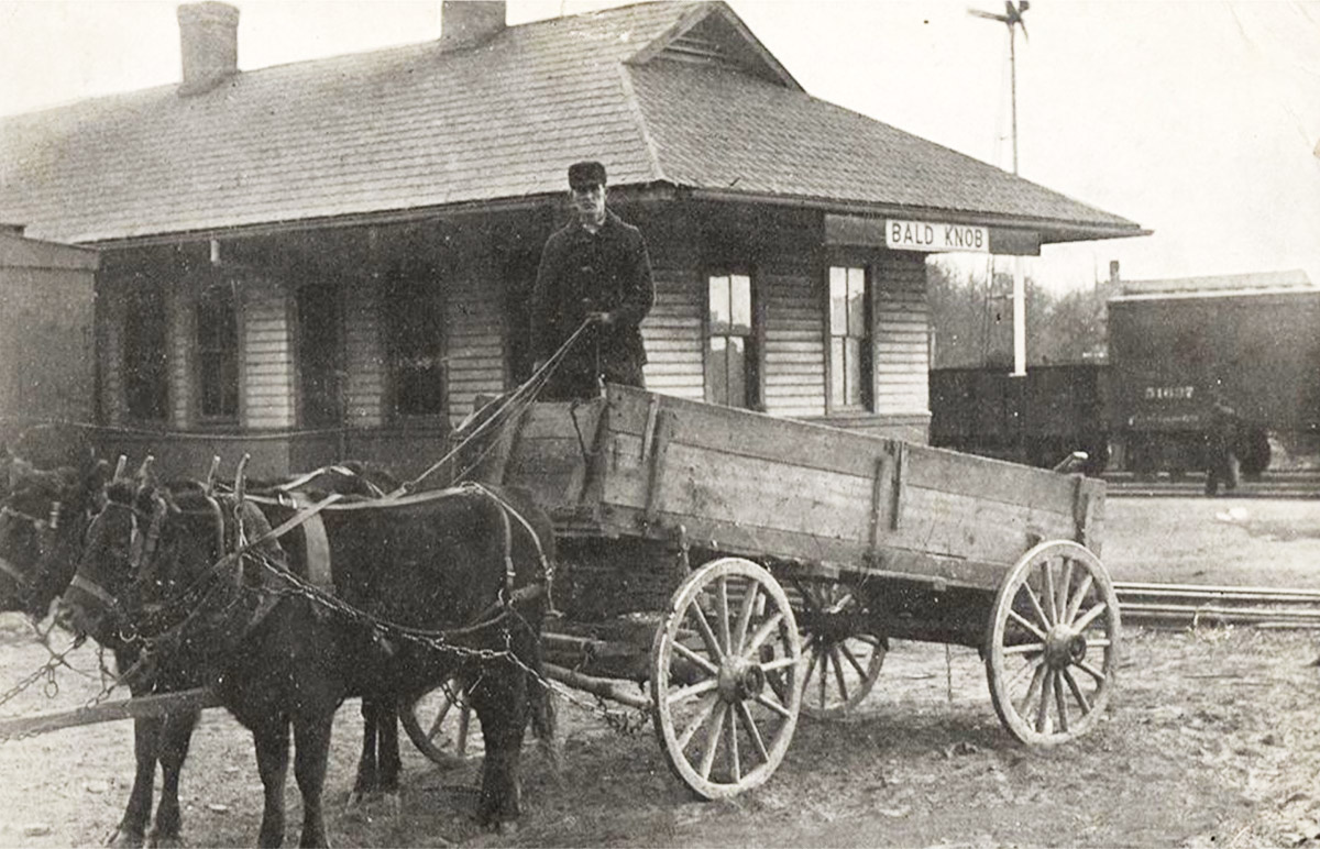 Man driving horses and wagon in front of single story building
