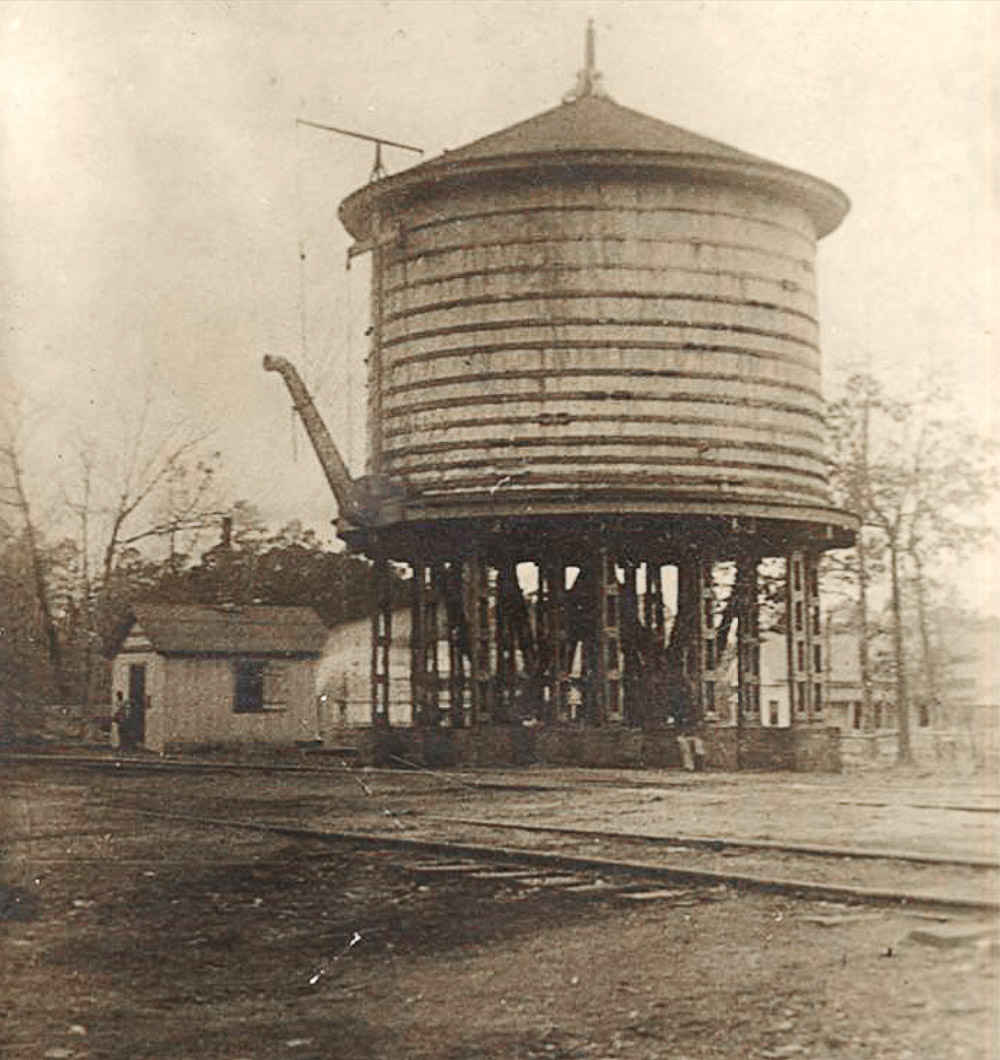 Single story wooden building beside large wooden water tank beside railroad tracks