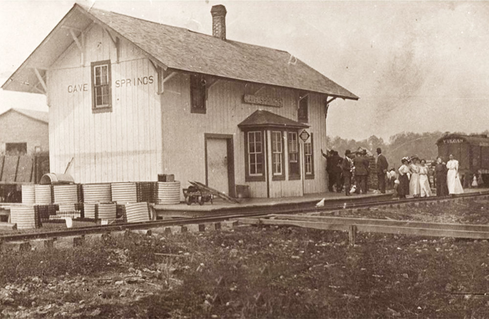 Single story wooden building beside railroad tracks "Cave Springs" with people standing on platform