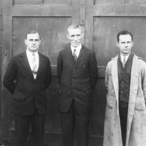 Three white men dressed in suits and ties standing before wooden door