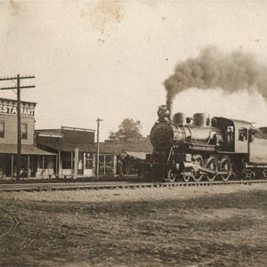 Train billowing smoke on tracks in front of row of wooden buildings