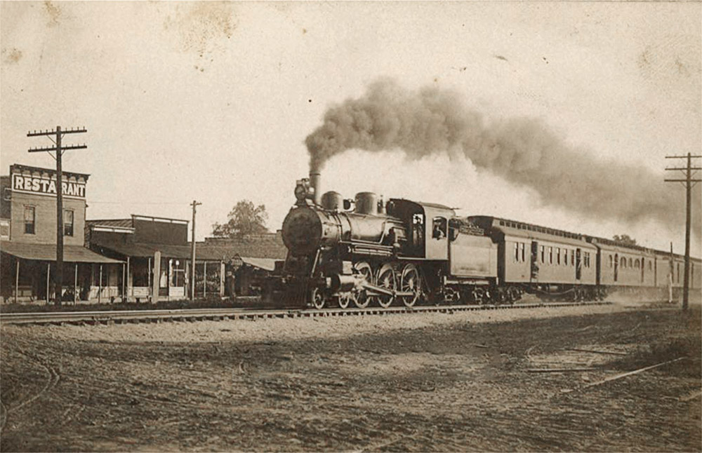 Train billowing smoke on tracks in front of row of wooden buildings