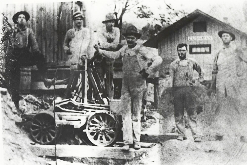 Six white men standing alongside machinery before single story wooden building