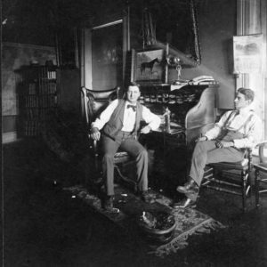 Two white men sitting in darkened office with a desk and paintings on the wall