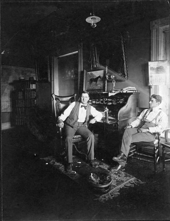 Two white men sitting in darkened office with a desk and paintings on the wall