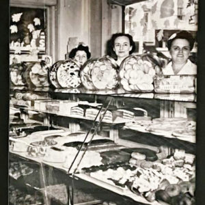 Three white women standing behind glass display case filled with baked goods